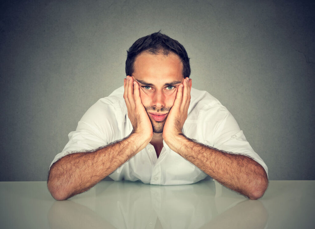 A man sits at a table, resting his face in his hands, appearing deep in thought or contemplation.